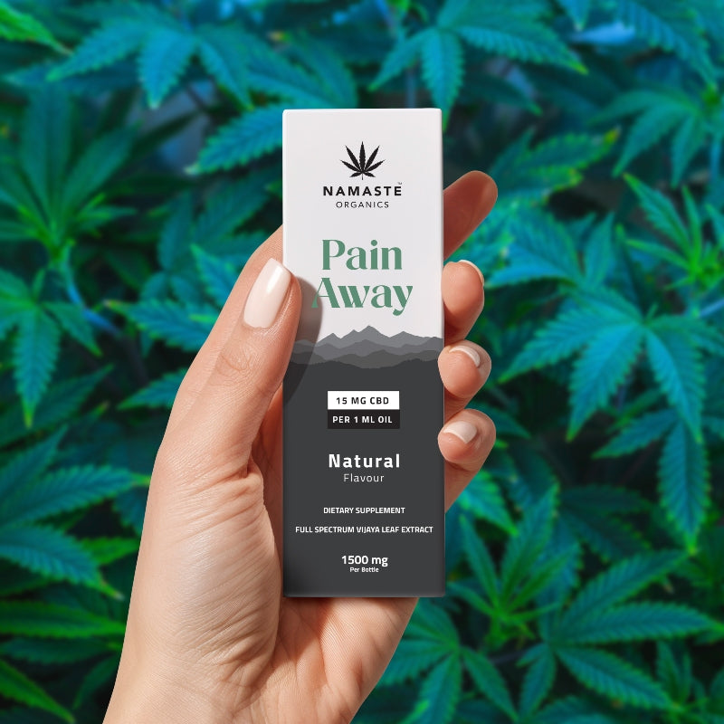 Hand holding a Namaste Organics Pain Away Full spectrum CBD Oil supplement box with cannabis leaves in the background