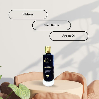 Navy blue bottle of conditioner with a gold cap on a beige pedestal. Labels "Hibiscus," "Shea Butter," and "Argan Oil" are visible.

