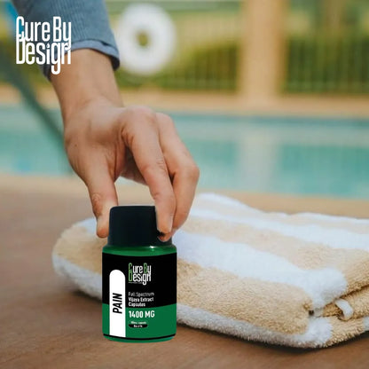 Hand holding a bottle labeled 'Cure By Design Pain Vijaya Extract Capsules' near a poolside with a towel.
