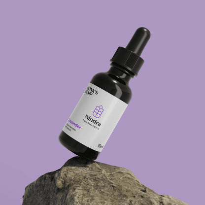 A dark glass bottle with a dropper cap labeled "Nindra Deep Sleep CBD Oil" balanced on a rock.