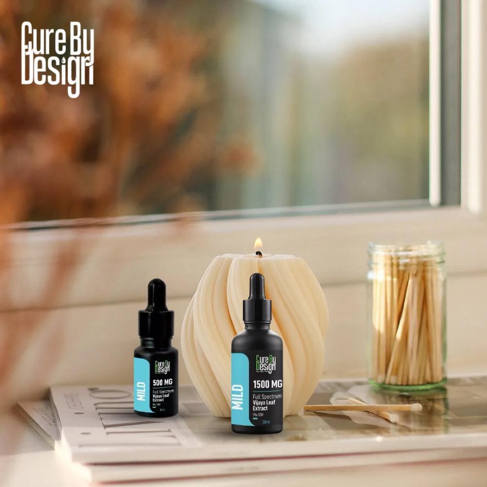 Two bottles of Cure By Design CBD Oil product on a surface with a candle and matches in the background.