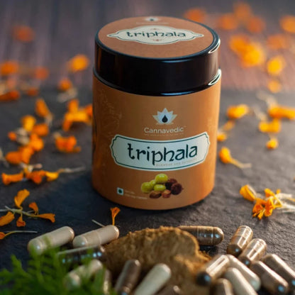 Jar of Cannavedic Triphala powder with capsules and flower petals on a dark surface.


